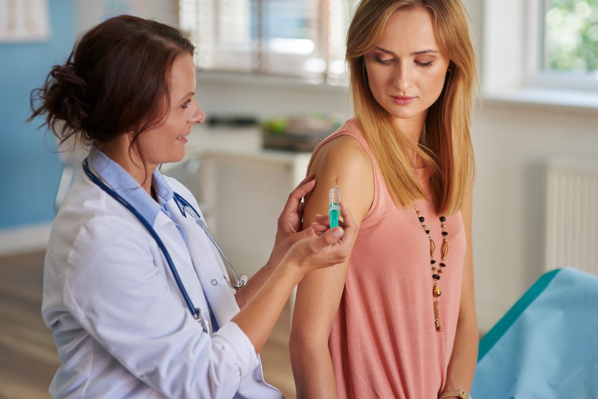 Woman in a doctors office getting a shot to the arm from her doctor. She is looking down at her arm as her doctor looks at the her with her thumb over the plunger.