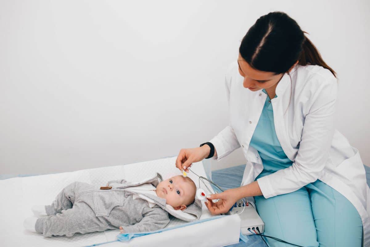 Baby in a onsie on a table in a medical setting. A doctor leans over the baby in the act of placing electrodes on its temples. 