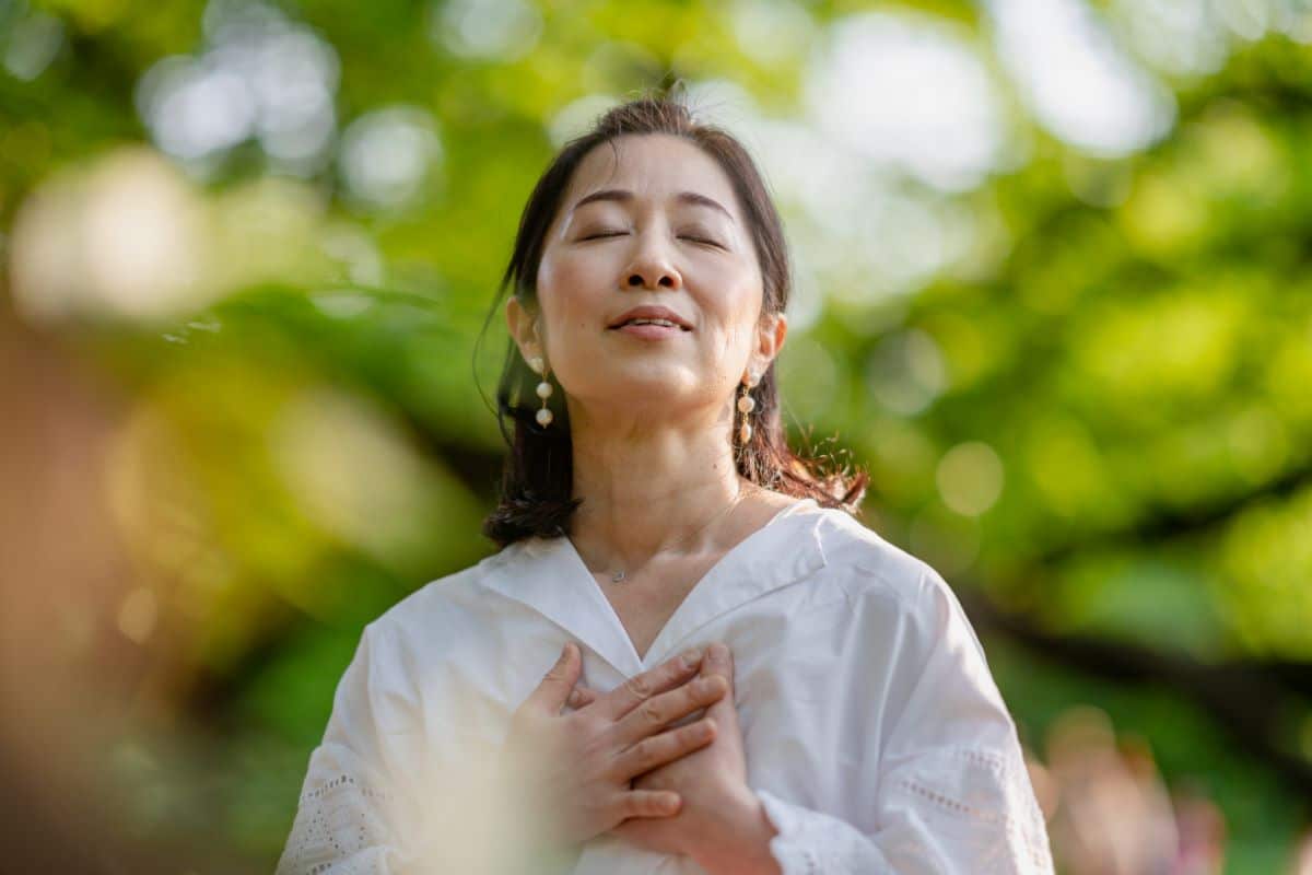 Woman in a park. Upper body and head are in frame. Her hands are clasping her chest. Her head is tilted slightly back and her mouth is barely open. Her eyes are closed in a look of joyful contemplation. The background is very blurry but includes the shape of trees and people.