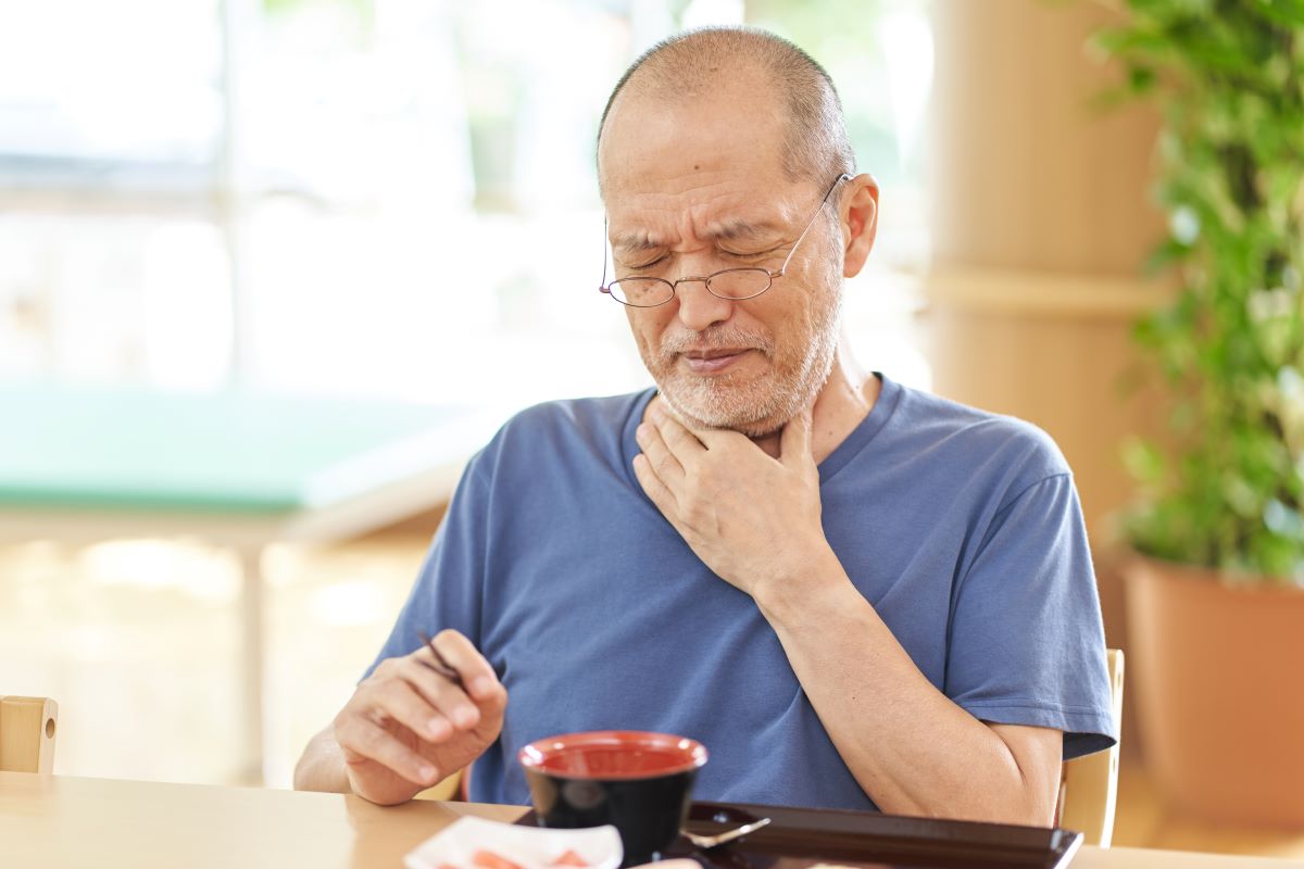 Older man at home closing eyes in pain with his hand placed on his upper chest and bottom of throat