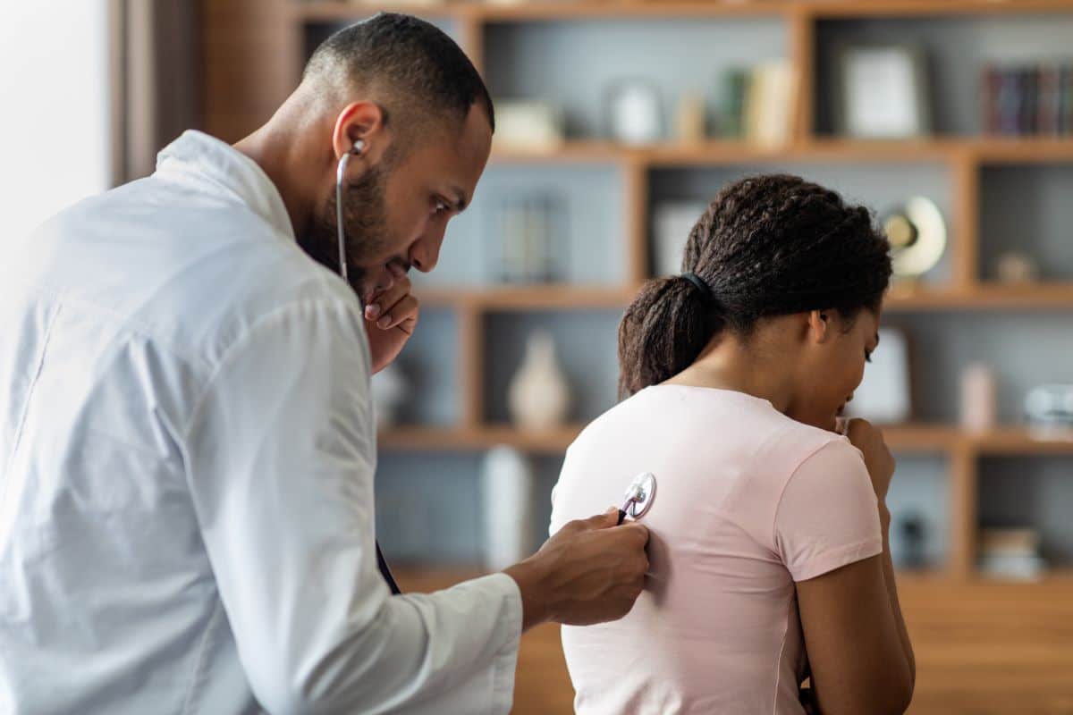 Male doctor in an office setting holding a stethoscope to a female patients back to listen to their lungs