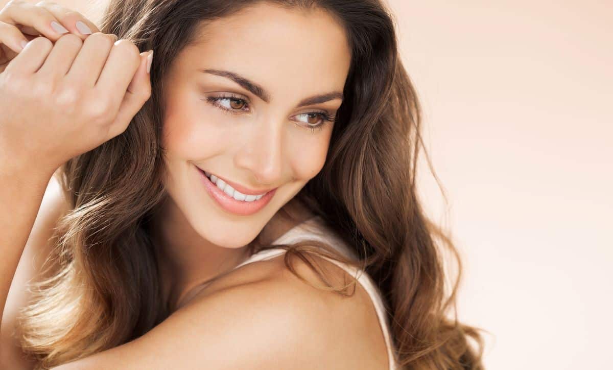 Closeup of brunette woman with hazel eyes smiling and looking towards the right while touching her hair. The background is beige. 