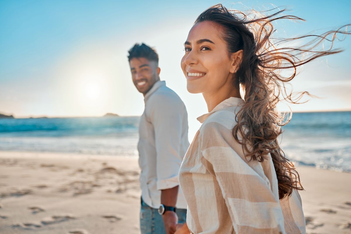 A heterosexual couple holding hands and walking on the beach as the wind blows the womans hair behind her. It is after midday and the ocean can be seen behind the couple. 