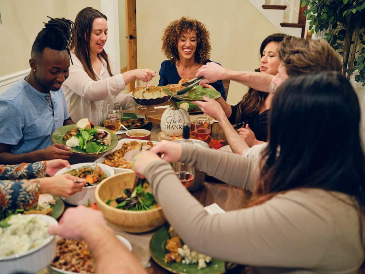 Multi-ethnic group of friends are sharing a large dinner in a home dining area that includes mashed potatoes, stuffing, and salad. They are all smiling or mid laugh.